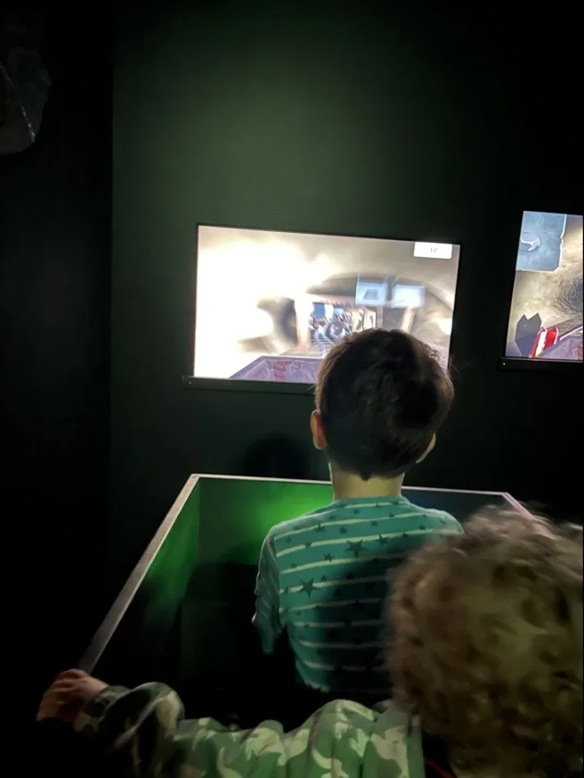 Zwei Kinder im Modell einer Lore vor einem Bildschirm vor der virtuellen Lorenfahrt