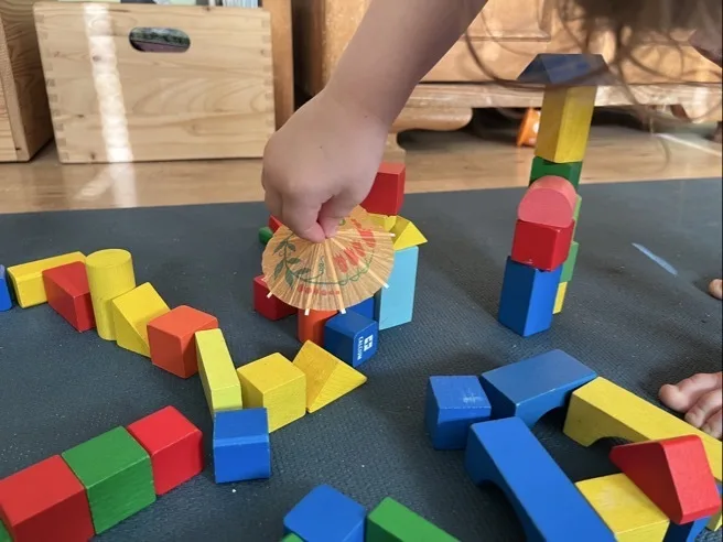 Bau aus Bauklötzen, dem gerade von einer Kinderhand ein Cocktailschirmchen hinzugefügt wird