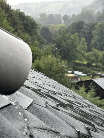 Regenwasser läuft aus einem Regenrohr aufs Dach, im Hintergrund Regendunst über Mittelgebirgslandschaft