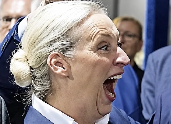 Profilaufnahme einer Person mit zurückgekämmtem, weißem Haar in einem Dutt; der Mund weit geöffnet, Zähne und Zunge sichtbar. Die Person trägt ein helles Hemd und ein dunkles Jackett; im unscharfen Hintergrund stehen mehrere weitere Personen.