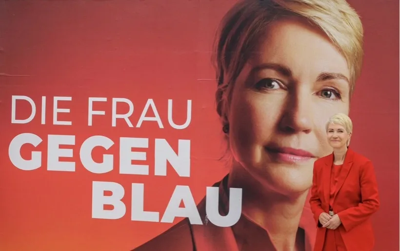 Großes rotes Plakat mit einer Nahaufnahme eines weiblichen Gesichts auf der rechten Seite und weißer Schrift links mit den Worten „DIE FRAU GEGEN BLAU“. Vor dem Plakat steht rechts eine Frau in einem roten Sakko und roter Hose, die die Hände vor dem Bauch gefaltet hat.