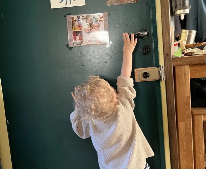 Ein kleines Kind mit blonden, lockigen Haaren ist von hinten zu sehen und steht an einer dunkelgrünen Tür, wobei die rechte Hand den schwarzen Türgriff erreicht. Auf der Tür sind vier Fotoabzüge befestigt; rechts neben dem Türrahmen sind ein Schloss und ein Teil einer Holzkommode mit Gegenständen sichtbar.