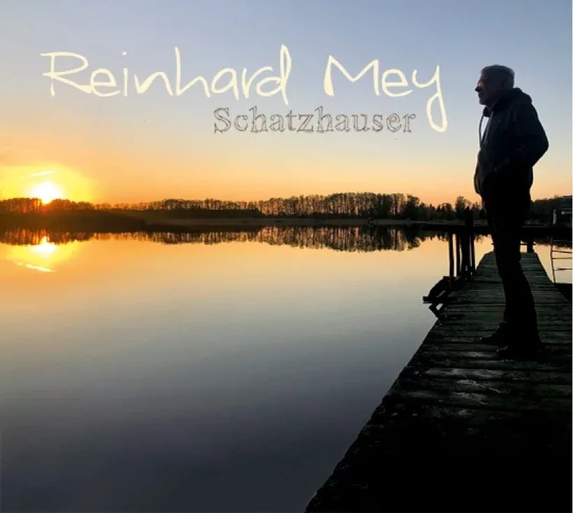 Sonnenuntergang über einem ruhigen See mit spiegelglatter Wasseroberfläche und einer niedrigen Baumreihe am Horizont. Auf einem hölzernen Steg rechts steht die dunkle Silhouette einer Person; oben im Bild ist große handschriftähn