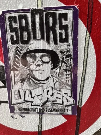 Auf einer strukturierten, rot-weiß lackierten Fläche ist ein rechteckiger Aufkleber mit lila Rand angebracht. Darauf ein schwarz-weißes Portrait einer Person mit Hut, Sonnenbrille und Zigarette, darüber große Blockbuchstaben „SBIURS“ und unten der Text „FREUNDSCHAFT UND ZUSAMMENHALT“.