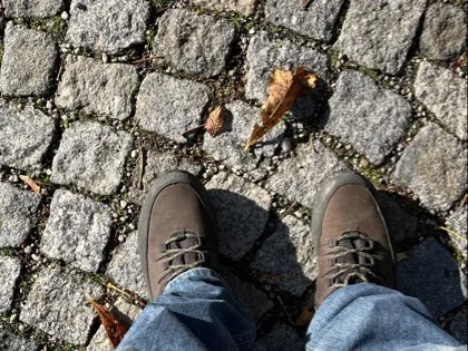 Braune Schnürschuhe in blauen Jeans stehen auf einem grauen Kopfsteinpflaster. Mehrere braune Herbstblätter und Schatten sind auf dem Pflaster sichtbar.