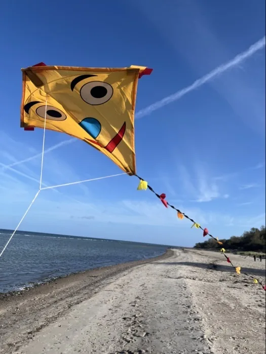 Gelbes, quadratisches Drachenpapier mit aufgemaltem Gesicht (zwei große Augen, blauer Nasenpunkt, roter Mund) und einer Schnur, an der eine bunte Wimpelkette hängt. Im Hintergrund sandiger Strand rechts, ruhiges Meer links und blauer Himmel mit Kondensstreifen.
