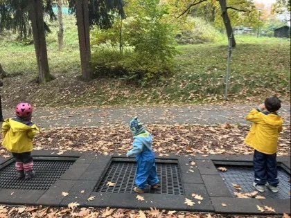 Drei Kinder in bunter Regenkleidung stehen mit dem Rücken zur Kamera auf nebeneinanderliegenden Metallgittern; das linke Kind trägt einen pinken Helm und gelbe Jacke, das mittlere blaue Latzhose und Mütze, das rechte eine gelbe Jacke und blaue Hosen. Im Hintergrund ein mit Herbstlaub bedeckter Weg, Rasenflächen und mehrere Bäume.
