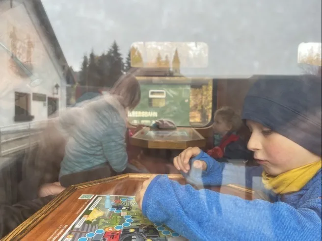 Ein Kind im Vordergrund rechts trägt eine dunkle Mütze, ein blaues Oberteil und einen gelben Schal und bewegt mit der rechten Hand Spielfiguren auf einem bunten Spielplan auf einem Holztisch. Durch das Fenster sind Reflexionen zu sehen; dahinter sitzen zwei weitere Personen an einem Tisch – eine mit hellblauer Jacke und ein Kind mit roter Jacke – und auf dem grünen Außenwagen ist halb das weiße Wort "BAHN" sichtbar.