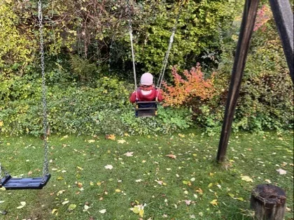 Ein Kind von hinten auf einer Schaukel, trägt eine rosa Mütze und eine rote Jacke. Links eine weitere leere Schaukel, Bodendecke aus Gras mit verstreuten Herbstblättern und im Hintergrund dichte Büsche mit grünen und orangen Blättern.
