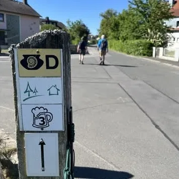 Wanderweg Hinweisschild mit schäumendem Bierglas im Vordergrund, im Hintergrund zwei Wanderer