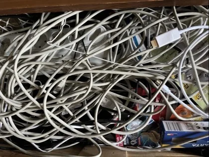 Verdrehte, ineinander liegende Kabel und Leitungen in Weiß, Grau und Schwarz füllen fast das gesamte Bild. Rechts unten liegen mehrere kleine, bunte Verpackungen und rechteckige Schachteln sowie ein sichtbarer weißer Steckeranschluss.