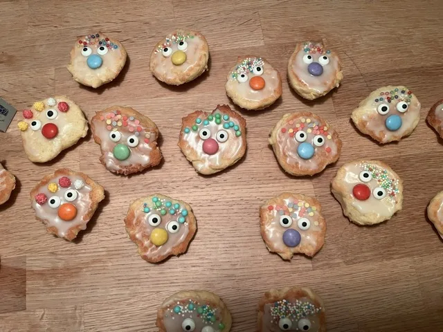 Mehrere kleine, unregelmäßig runde Kekse mit weißer Zuckerglasur liegen auf einer Holzoberfläche. Jeder Keks trägt zwei runde Zuckeraugen, eine einzelne bunte Schokolinsen‑Dekoration in der Mitte und verschiedenfarbige Zuckerstreusel obenauf.