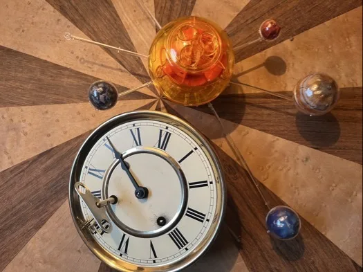 Rundes Zifferblatt mit römischen Zahlen, zwei schwarzen Zeigern und einem Metallaufziehschlüssel auf der Oberfläche. Daneben eine transparente orangefarbene Kunststoffkugel mit sichtbaren Zahnrädern und mehrere kleine farbige Kugeln (blau, rot, grau) an dünnen Metallstäben auf einem holzgemusterten Untergrund.