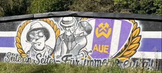 Wandgemälde in den Farben Lila, Weiß, Gelb mit links einem Portrait einer Person mit Hut, Sonnenbrille und Zigarette, in der Mitte zwei gekleidete Personen im Kuss bzw. enger Umarmung und rechts einem Schild mit gekreuztem Hammer und Bergbausymbol sowie den Buchstaben „AUE“, umgeben von Lorbeerzweigen. Am unteren Bildrand ist in geschwungener Schrift ein Slogan zu lesen, teilweise von Gras bedeckt.