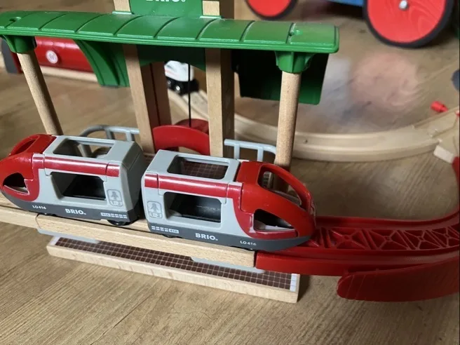 Roter und hellgrauer BRIO-Spielzeugeisenbahnwagen mit zwei Waggons steht auf hellbraunen Holzschienen. Darüber ein grünes Bahnhofsüberdach mit vier runden Holzsäulen; im Hintergrund ein rotes Rad und weitere Schienenstücke auf einem Holzfußboden.
