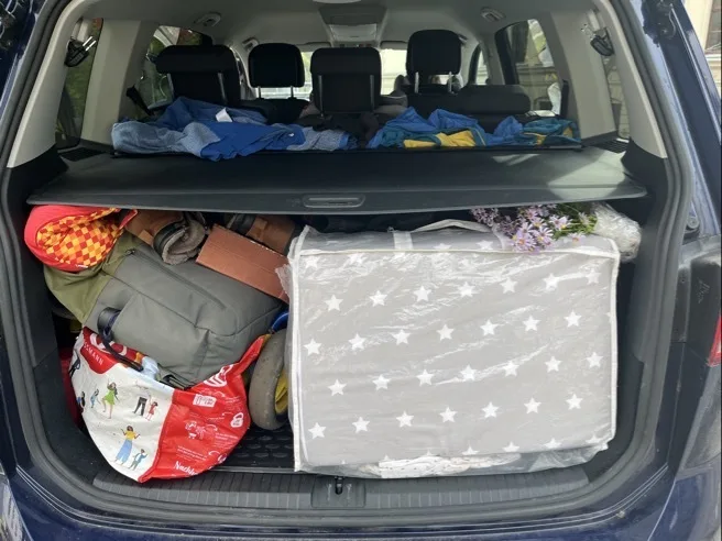 Offener Kofferraum eines dunklen Fahrzeugs, im rechten Bereich ein grauer, plastikverpackter Kasten oder Karton mit weißen Sternen, darüber eine durchsichtige Folie mit kleinen lila Blumen. Linke Seite gefüllt mit einer roten Einkaufstasche mit bunten Figuren, einem grünen Rucksack, einem braunen Gürtel und weiteren Taschen; in der oberen Ablage liegen mehrere gefaltete Kleidungsstücke, drei Kopfstützen sind sichtbar.