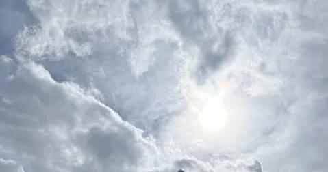 Himmel mit weißen Wolken, durch die die Sonne als Runde, helle Lichtquelle zu sehen ist