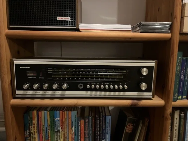 Holzkasten mit mehreren Regalböden, auf dem mittleren Regal ein silber-schwarzes Stereo-Radio mit Skala, Knöpfen und Drehreglern. Auf dem unteren Regal Vorderkanten vieler bunt gebundener Bücher, auf dem oberen Regal Stapel flacher Hefte und CDs.