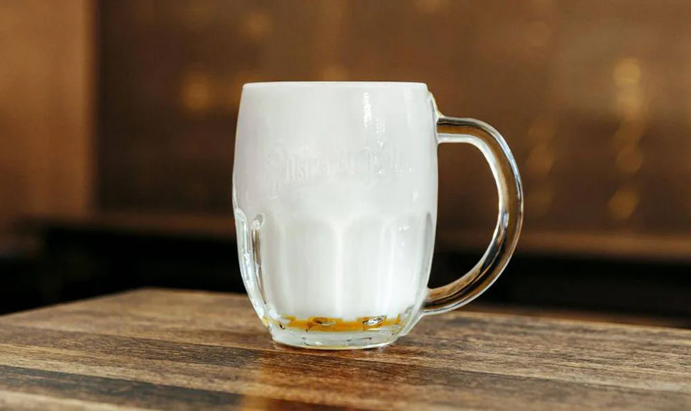 Ein transparenter Glasbierkrug mit Griff steht auf einer dunklen Holzoberfläche. Im Krug befindet sich eine weiße Schaumschicht und ein kleiner Rest goldbrauner Flüssigkeit am Boden; der Hintergrund ist unscharf und bräunlich.