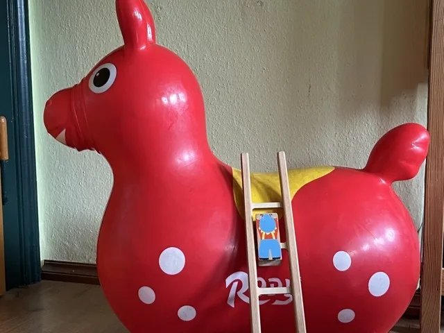Rotes, aufblasbares Tier mit großem weißen Auge, mehreren weißen Punkten, gelber Fläche auf dem Rücken und kurzem erhobenem Schwanz. Eine kleine Holzleiter lehnt an der Seite, zwischen den Sprossen steht eine bemalte Holzfigur; heller Boden und beige Wand im Hintergrund.