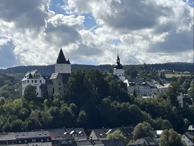 Auf einem bewaldeten Hügel stehen mehrere historische Gebäude mit Turm- und Schaftdächern, darunter ein weißer Turm mit spitzem Dach und ein Kirchturm mit Zwiebelhaube. Im Vordergrund Reihenhäuser mit dunklen Dächern; darüber großer Himmel mit dichten, teils aufgewühlten Wolken.