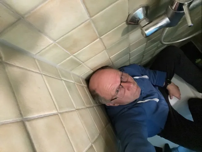 Ein Mann mit Brille und kahlem Oberkopf trägt einen blauen Kapuzenpullover und sitzt in einer weißen Duschtasse. Beige quadratische Wandfliesen und eine verchromte Duscharmatur mit Handbrause sind links und oberhalb zu sehen; die Person blickt zur Kameralinse.