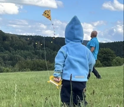 Ein kleines Kind von hinten in einer hellblauen Kapuzenjacke hält in der rechten Hand einen gelben Drachengriff; der Drachen mit gelbem Gesicht fliegt an einer Schnur im blauen Himmel. Im Hintergrund steht weiter entfernt ein erwachsener Mann in türkisblauem Oberteil auf einer grünen Wiese vor einem bewaldeten Hügel unter wolkigem Himmel.