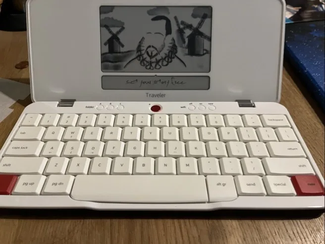 Weißes, klappbares Portable-Keyboard mit hellen, quadratischen Tasten und zwei roten Tastecken auf einer Holzoberfläche. Obere Klappenseite zeigt ein Graustufen-Display mit einer skizzenhaften Darstellung von zwei Windmühlen und einer Person sowie einer handschriftlichen Zeile unter dem Display; auf dem Gehäuse ist das Wort „Traveler“ zu lesen.