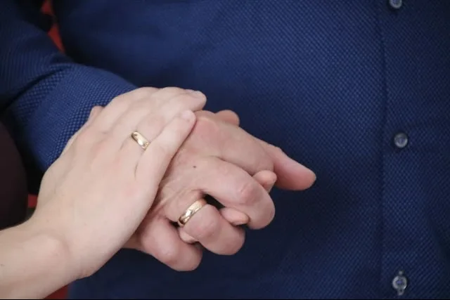 Zwei Hände, die sich halten, eine dritte Hand darüber, zwei Eheringe