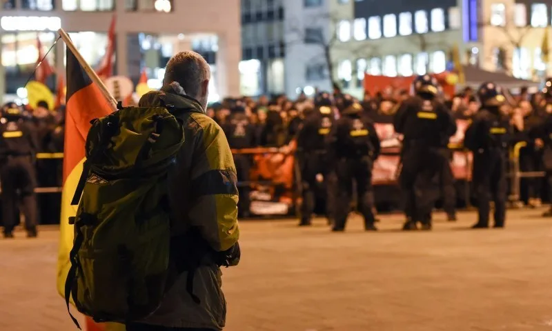 Mann von hinten mit schwarz-rot-goldener Fahne blickt auf eine Hunderschaft Polizisten in Sturm-Montur, die ihm den Rücken zuwenden